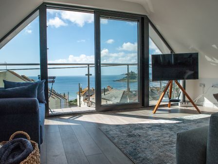A living room with a dark blue sofa a wooden tripod TV stand a rug and large windows overlooking the sea at Marazion Attic in Marazion