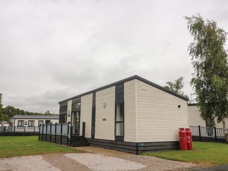 A single story modular home with a ramp and stairs on a grassy plot with two red propane tanks and trees nearby at Waterside Lodge South Lakeland Leisure Village near Carnforth