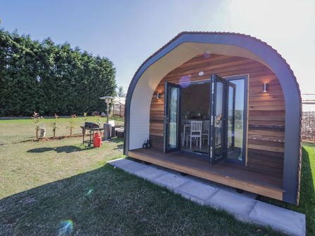 An arched wooden cabin with open glass doors on a grassy lawn with patio heaters and a barbecue grill at Tamina in Hillhampton Great Witley