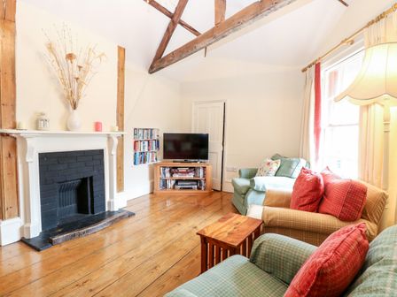 A living room with a fireplace a television on a stand shelves with books and games two sofas with cushions a wooden floor and exposed ceiling beams at Forge House in Cley Next The Sea