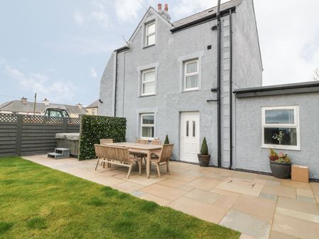 A patio with wooden table and chairs next to a gray house and a green lawn at Fishermans Cottage in Malltraeth