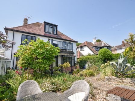 A garden with a round glass table and wicker chairs outside houses with tiled roofs and black window frames at Coram Cottage in Lyme Regis