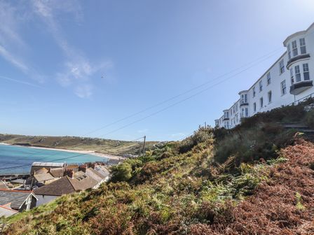 A hillside with vegetation and white buildings overlooking a beach and ocean at Starfish in Sennen Cove
