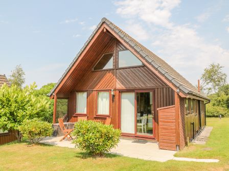 A wooden cabin with a sloped roof a patio with chairs and bushes in the yard at Lodge 16 in Lanteglos near Camelford