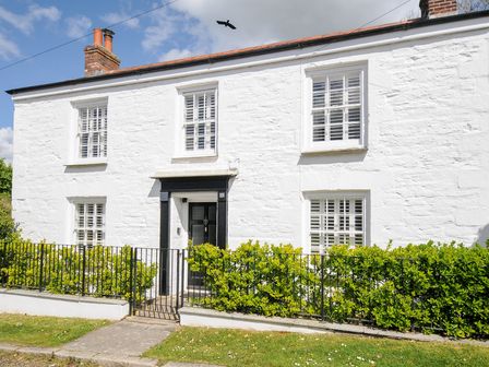 The front exterior of a white stone house with a black door white window shutters and a garden with shrubs behind a black metal fence at Quay House in Charlestown