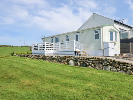 An exterior view of a single-story caravan with a white deck and stone retaining wall on a grassy landscape at Ty Newydd Caravan in Llannor near Pwllheli