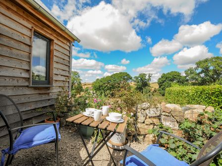 An outdoor seating area with two chairs with blue cushions and a small wooden table with cups and saucers near a wooden building and a stone wall with plants at Lillemor South Tawton near Whiddon Down