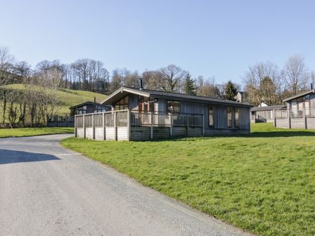 A wooden cabin with a fenced porch on a grassy plot near a gravel road at Rivendell Lodge in Roger Ground near Hawkshead