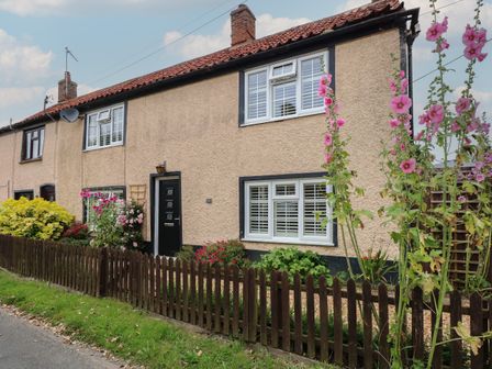 A beige terraced house with white windows a black door and flowers behind a wooden fence at Well Cottage in Grimston