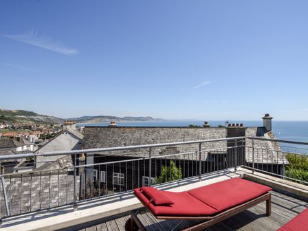 A rooftop terrace with a red lounge chair overlooking rooftops and the sea at 5 St Michaels House in Lyme Regis