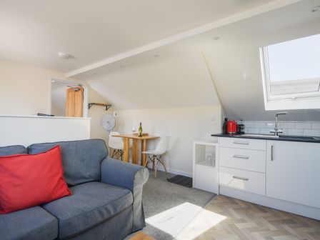 A living room with a gray sofa and red pillow a wooden table with white chairs a kitchen with white cabinets and a red kettle at Ayla Lyme Regis