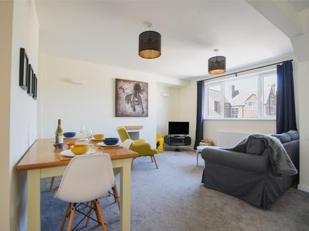 A living room with a dining table set with bowls and glasses a gray sofa a yellow armchair a TV and large windows with dark curtains at Bahari in Lyme Regis