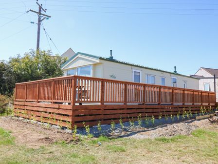 A beige caravan with multiple windows and a green roof on a grassy area with a wooden deck surrounding it at Gorphwysfa Caravan in Tregele near Cemaes Bay