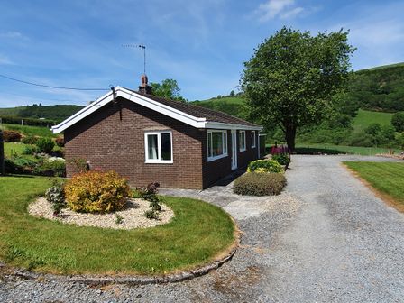 A brown brick house with white trim surrounded by grass and bushes with a gravel driveway at Llwynon in Goginan near Aberystwyth