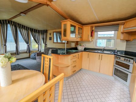 A kitchen and living area with wooden cabinets a round table with chairs a grey sofa and large windows with curtains at Rosehaven in Tegryn near Crymych