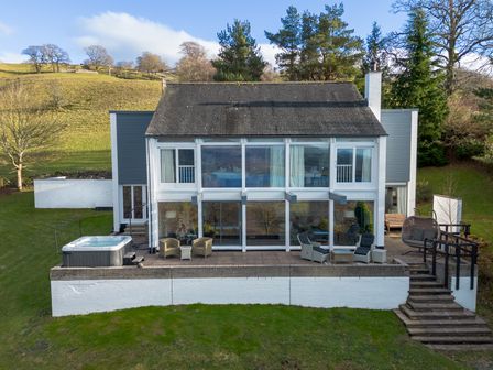 A two-story house with large windows a patio with chairs and a hot tub surrounded by grass and trees at Paddock House in Ambleside