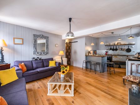 An open-plan living room and kitchen with purple sofas, yellow and orange cushions, a coffee table with sunflowers, bar stools at a kitchen island, and hanging pans at Pibbies in Port Isaac