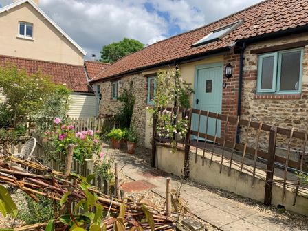 An exterior view of a stone and brick house with blue doors and windows and a tiled roof with plants and flowers along a path at The Old Dairy in Colyton