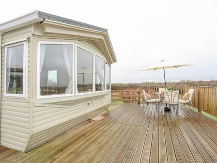 A deck with outdoor table chairs and umbrella next to a mobile home at Mountain View Lodge in Caergeiliog near Valley