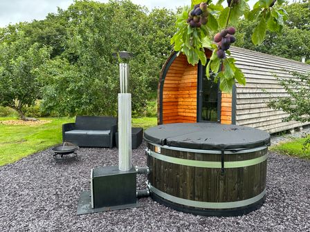 An outdoor area with a wooden pod, a round hot tub with a cover, a black sofa, a fire pit, and trees in the background at Tyddyn Parthle Pod 1 in Bontnewydd Gwynedd