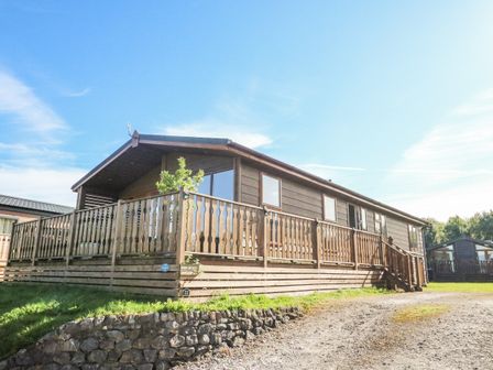 A wooden cabin with a fenced porch and stairs surrounded by grass and gravel at South Lakeland Leisure Village