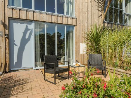 An outdoor patio with two black chairs and a table with tea set and croissants outside a wooden building with large windows at Mousehole in Mawnan Smith Near Falmouth