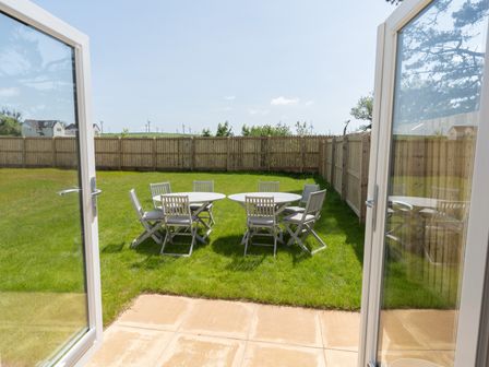 An outdoor patio area with two round tables and several chairs on green grass behind glass doors at The Bright House in St Eval near St Columb Major