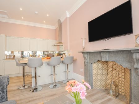 A kitchen with a breakfast bar and stools and a fireplace with a TV above it at The Stannary in Helston