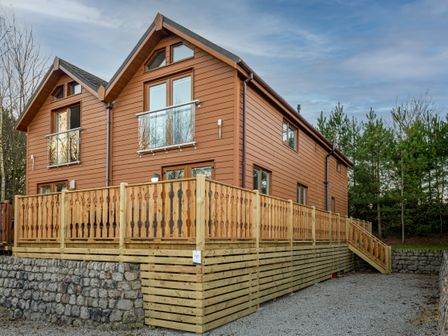 A wooden house with a raised wooden deck and stone retaining wall surrounded by trees at Lakeland Lodge in Carnforth