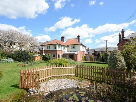 A garden with a wooden fence around a pond and a house with a tiled roof in the background at Holly Lodge in Aldeburgh
