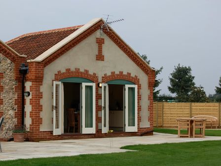 A single story building with two open French doors leading to an outdoor patio with wooden table and chairs at Romulus in Larling near East Harling