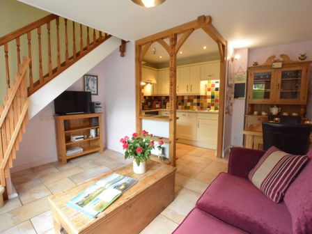 A living room with a purple sofa and wooden coffee table with flowers a wooden cabinet and a kitchen with tiled walls at Seafarers in Fressingfield