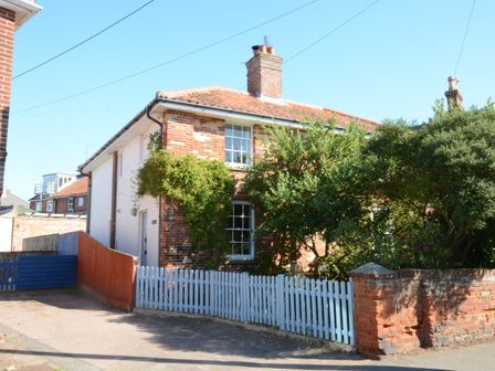 A brick house with a tiled roof surrounded by greenery and a light blue picket fence at Pebble Beach Cottage Aldeburgh