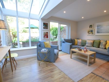 A living room with blue sofas yellow and green cushions a wooden coffee table and large windows at Seawall Cottage Aldeburgh