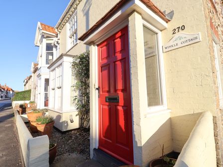 A street view of a house with a red door and beige walls displaying a sign reading Winkle Cottage at 270 in Aldeburgh