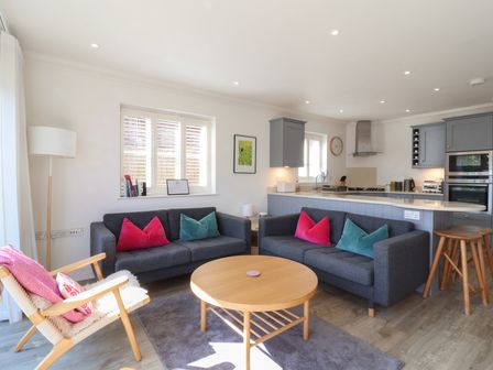 A living room with two gray sofas, a round wooden coffee table, a wooden armchair with pink cushions and an open kitchen with gray cabinets at Crabapple Cottage Southwold