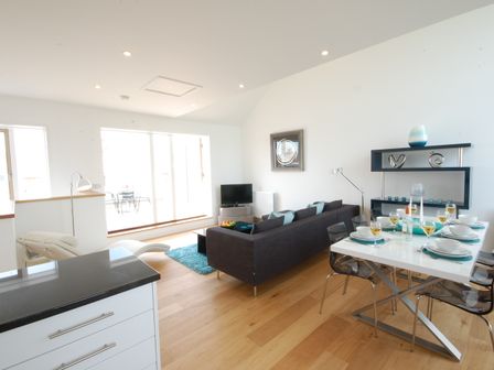 A living room and dining area with a dark sofa a white dining table set with glasses and plates and shelves with decorative items at Seal Cove in Southwold