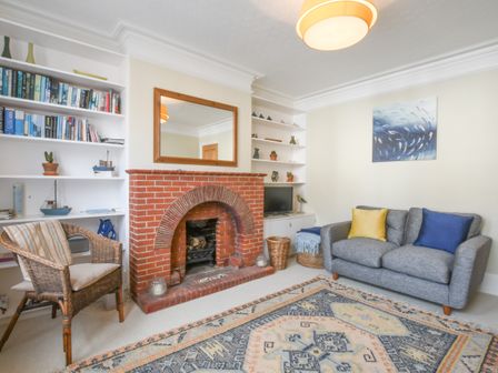 A living room with a brick fireplace a gray couch with blue and yellow cushions a wicker chair and built in shelves at Spindrift Southwold