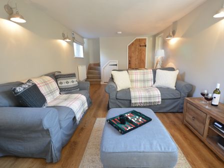 A living room with two sofas, an ottoman with a backgammon board, a wooden cabinet with wine glasses and a bottle of wine at Little Turnpike Cottage, Melton in Woodbridge