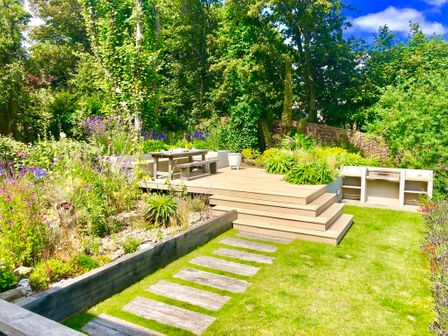 A garden with a wooden deck and outdoor table surrounded by plants and trees at Aldeburgh Lodge Gardens in Aldeburgh