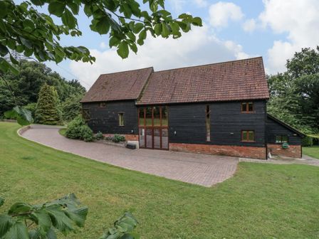 An exterior view of a barn-style building with black wooden siding and a tiled roof surrounded by grass and trees at Quill Farm Barn in Campsea Ashe