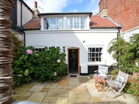 A white brick house with a tiled roof and two windows with an open door in the middle a paved courtyard with two wooden chairs a small table and plants at Mariners Cottage King Street in Aldeburgh