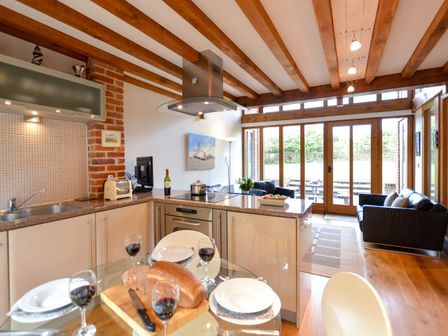 An open kitchen and living area with wooden beams glass doors and a dining table with bread and wine at Two Chantry Barns Orford