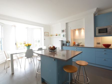 A kitchen with blue cabinets an island with stools a dining table with chairs and a vase of flowers at Moot Green House Aldeburgh