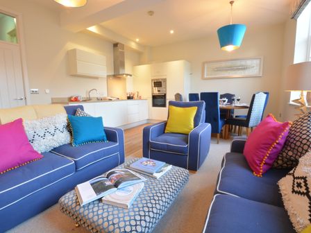 A living room with blue sofas and colorful cushions and a kitchen and dining area at Skimming Stones Aldeburgh in Aldeburgh