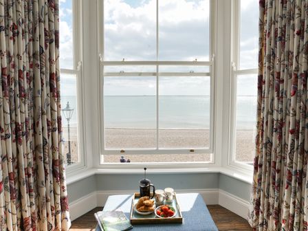 A bay window with floral curtains overlooking a pebble beach and sea with a tray of breakfast items on a blue ottoman at Shingle Skies Aldeburgh
