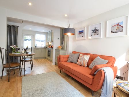 A living room with an orange sofa and three framed pictures above it next to a dining table with two chairs and a kitchen in the background at 53 Tern Cottage Aldeburgh