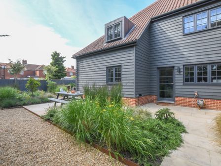 A garden with a gravel path and patio area with a picnic table next to a two-story house with gray siding and red brick at Corner Cottage Thorpeness
