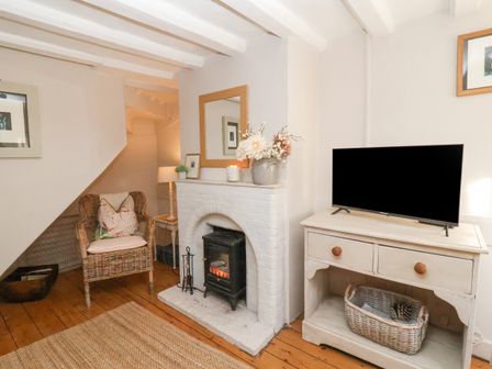 A room with a TV on a wooden cabinet a fireplace with flowers and a mirror above a wicker chair under stairs at Rosemary Cottage Aldeburgh