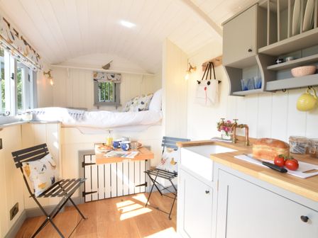 A compact kitchen and dining area with a small wooden table set between two black folding chairs and a raised bed in the background at Shepherds Pightle in Hollesley